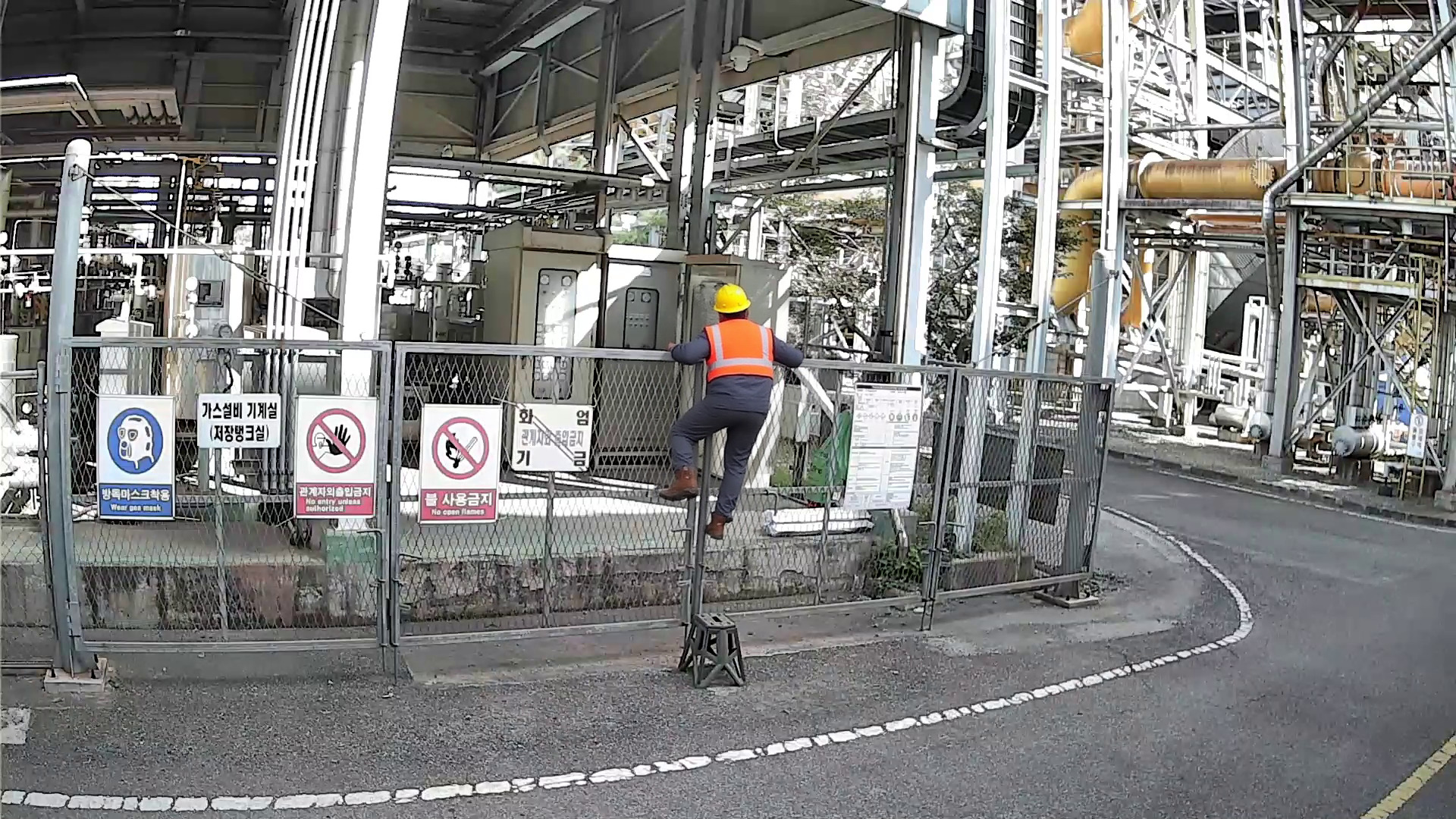 Male worker in a yellow safety helmet, orange high-visibility vest over a dark long-sleeve shirt, gray work trousers, brown safety boots